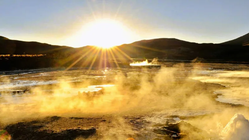 Géiseres del Tatio al amanecer con vapor dorado iluminado por el sol naciente y montañas al fondo
