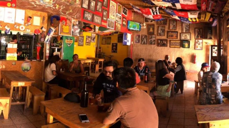 Interior de bar animado com pessoas sentadas em mesas de madeira, paredes decoradas com fotos, bandeiras e escudos de times
