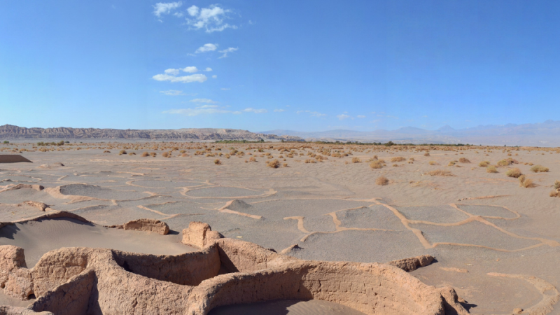 Ruinas de adobe de la Aldea Tulor en el desierto de Atacama, con suelo agrietado, vegetación escasa y montañas al fondo.