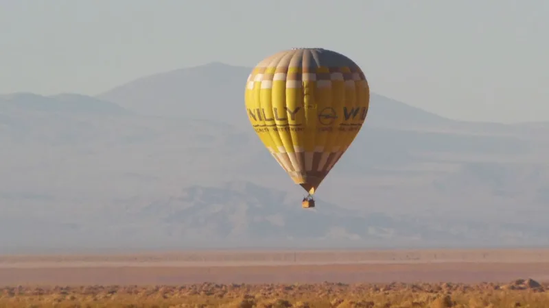 Globo aerostático amarillo sobrevolando el desierto de Atacama con montañas al fondo bajo un cielo brumoso.