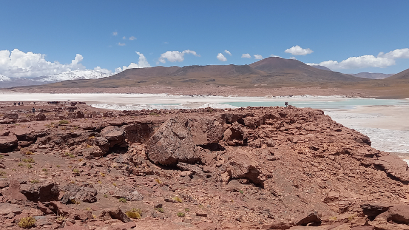Formaciones de rocas rojas en el Mirador Piedras Rojas, con laguna turquesa, salar blanco y volcanes nevados al fondo.
