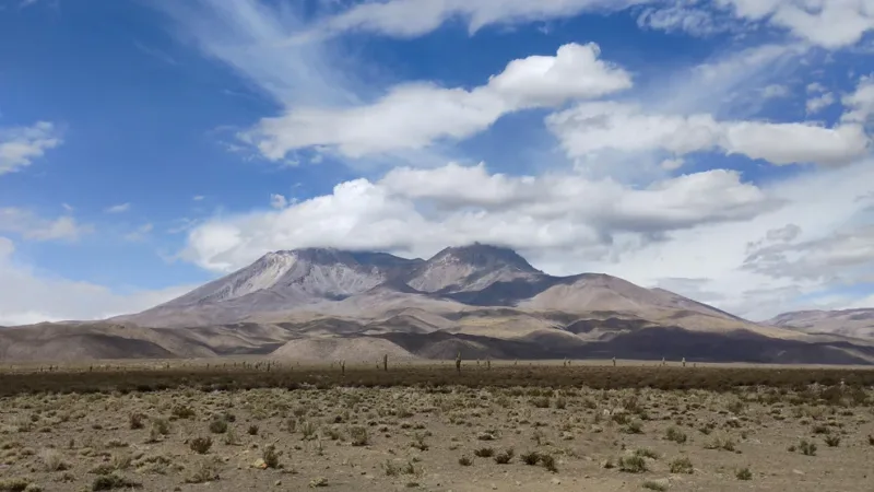 Vast arid Atacama Desert landscape with sparse scrub, rolling hills, and a volcanic mountain peak under a blue cloudy sky