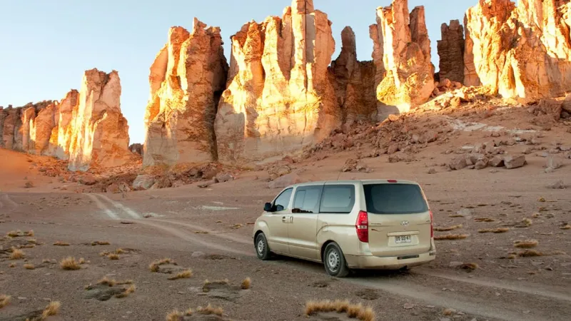 A van on a dirt track in the Atacama Desert, with towering golden sandstone rock formations glowing in warm sunlight.