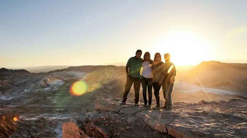 Four people posing on a rocky ridge in the Atacama Desert at sunset, with dramatic landscape and lens flare behind them