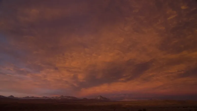Dramatic sunset over the Atacama Desert with fiery orange and purple clouds above a vast arid plain and distant volcanic