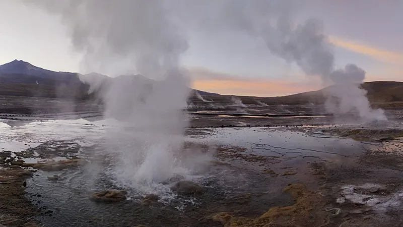 Steaming geysers erupting at dawn in the Atacama Desert, with volcanic mountains and a colorful twilight sky in the