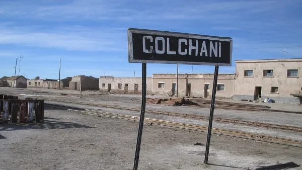 Placa com o nome "Colchani" em estação ferroviária abandonada, com trilhos e construções antigas ao fundo.