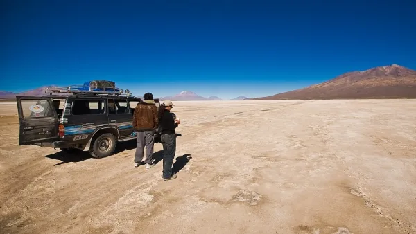Duas pessoas ao lado de um jipe 4x4 no árido Deserto de Chiguana, com vulcões ao fundo e céu azul intenso.